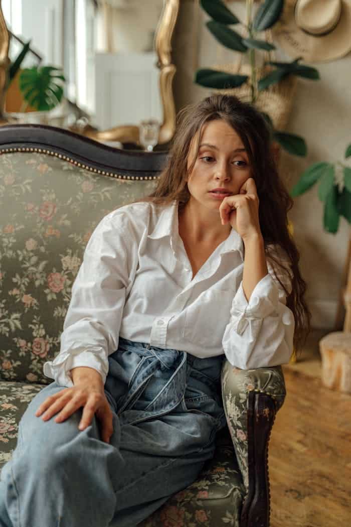 A woman in a casual outfit sits thoughtfully on a floral vintage sofa indoors.
